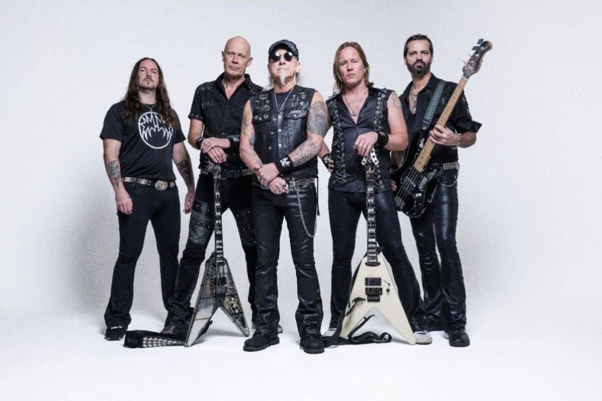 Five male rock band members pose together in black outfits with guitars against a white studio background.