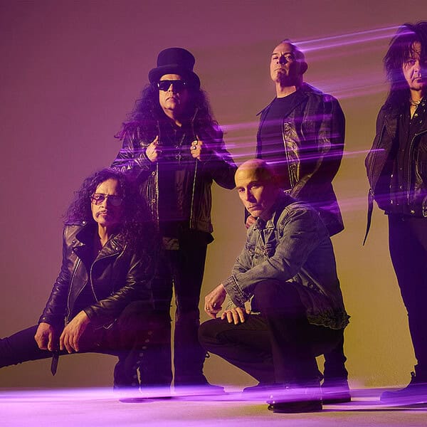 Promo shot of a rock band posing in a studio, five members wearing leather jackets and dark clothing with purple light streaks in the background.