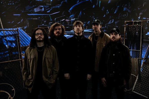 Five-member rock band posing in a dim, industrial backstage area with blue lighting.