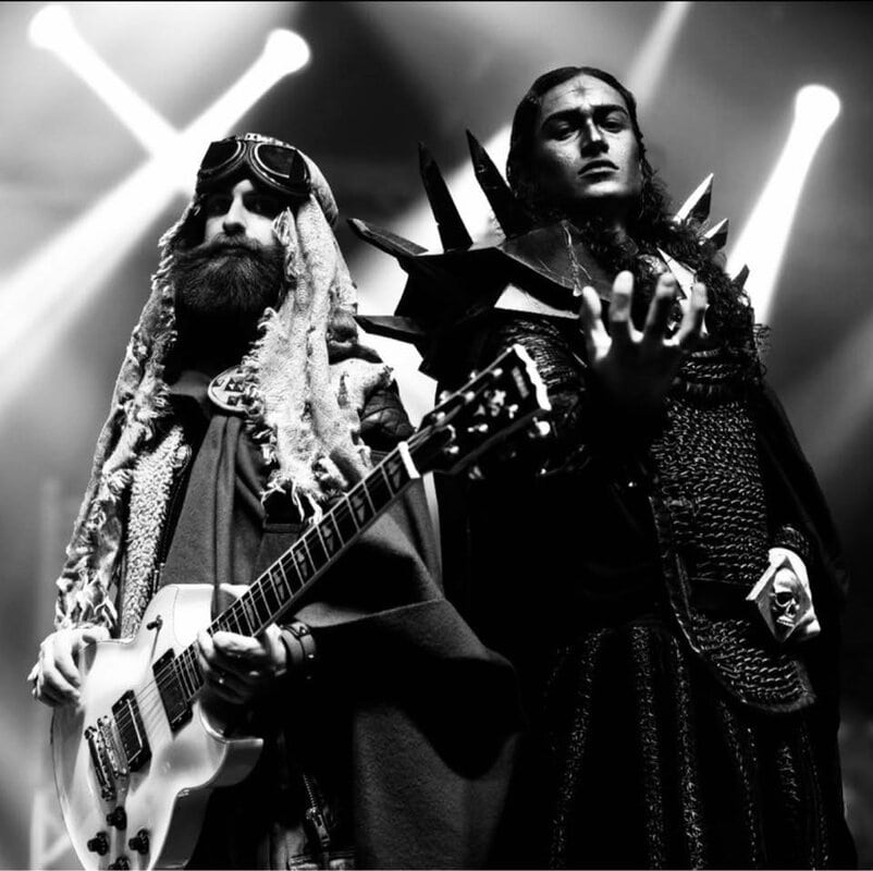 Two performers on stage in elaborate costumes; one guitarist with a white guitar, the other in spiked armor under bright lights (black and white photo).