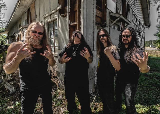 Four male rock/metal musicians in black shirts pose outside a dilapidated wooden building, flashing metal horns toward the camera.