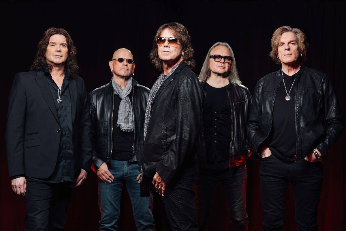 Five male rock musicians in black outfits pose together on a dark stage, some wearing sunglasses and leather jackets.