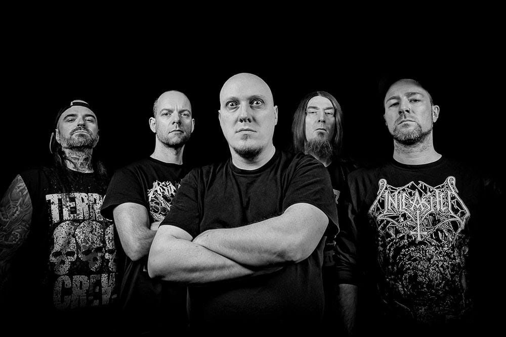 Five male metal band members stand against a black background, wearing dark T-shirts and looking stern. The central figure has his arms crossed.