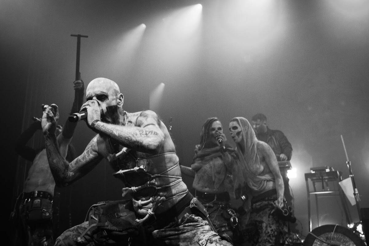Black-and-white live concert: a bald, tattooed vocalist sings into a microphone, surrounded by performers in skull makeup behind him in a dark stage. NOTE: concise alt text for informative image, not overly long.