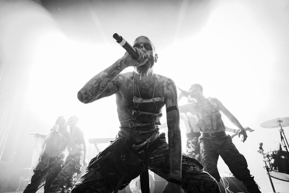 Lead singer with tattoos and chest harness sings into a microphone on a metal concert stage, band members and drums visible in the background under bright lights.
