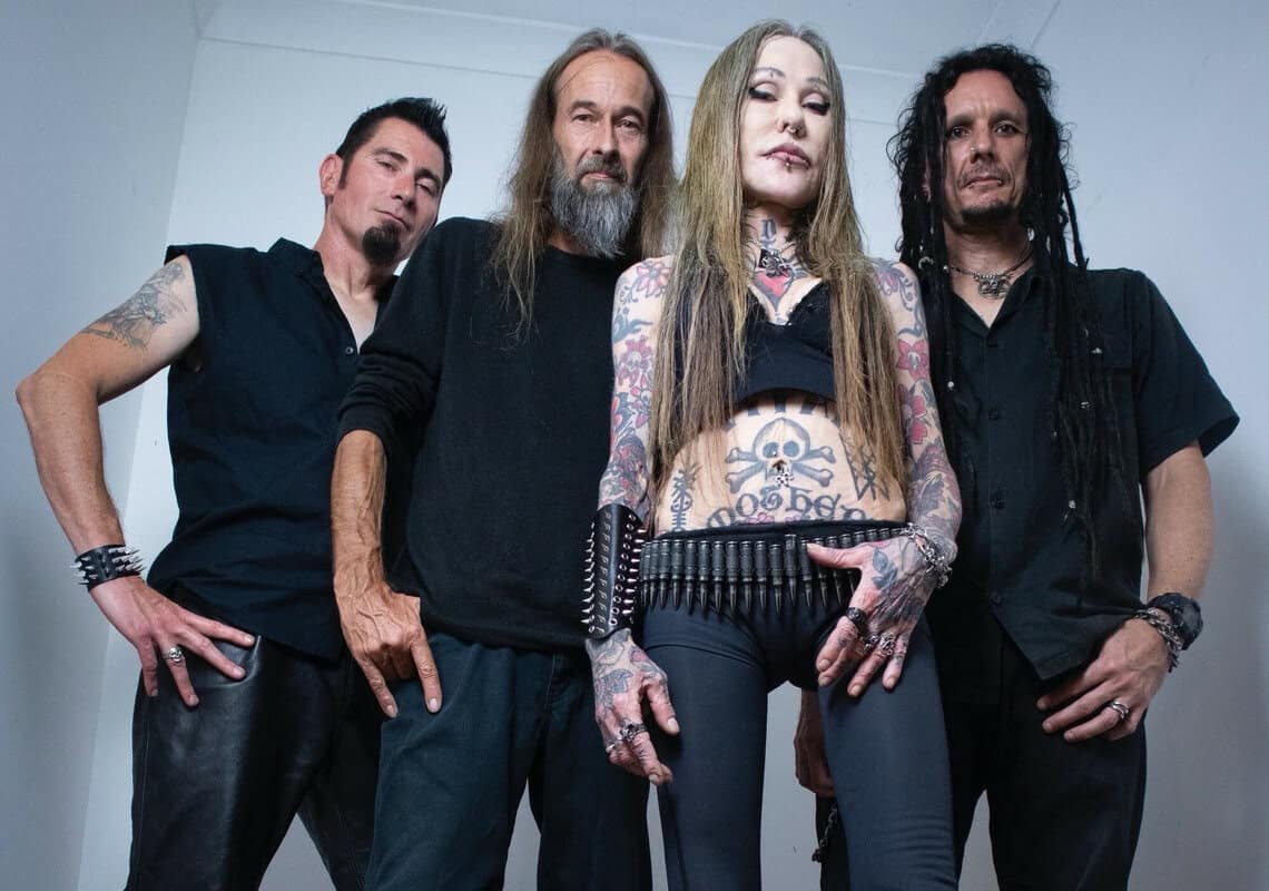 Tattooed female rocker at center wearing a studded belt, flanked by three men in black in a studio.