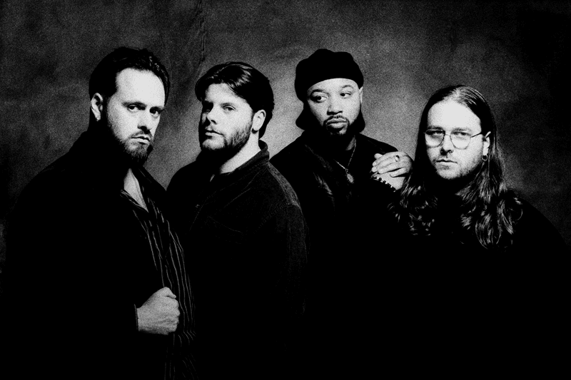 Four men stand together in a dark studio, posing for a band portrait with serious expressions.