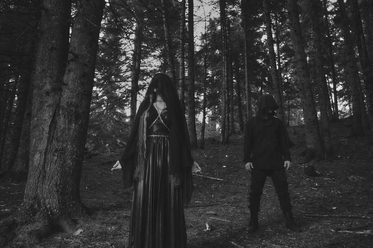 Two hooded figures stand in a dark forest; a woman in a long black cloak with a veiled face stands with arms outstretched, a man in dark clothing nearby.