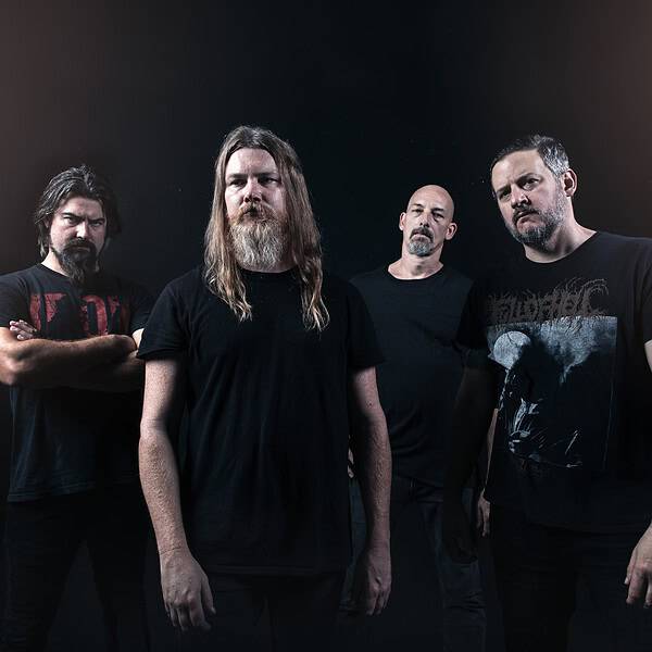 Four men in black T-shirts pose in a dim studio, forming a metal band portrait.