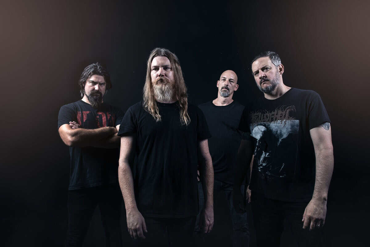 Four men in black T-shirts pose in a dim studio, forming a metal band portrait.