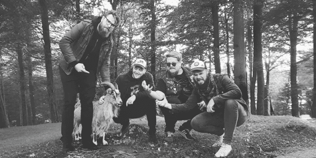 Four men with a scruffy dog posing in a forest, some crouching and others standing, smiling at the camera.