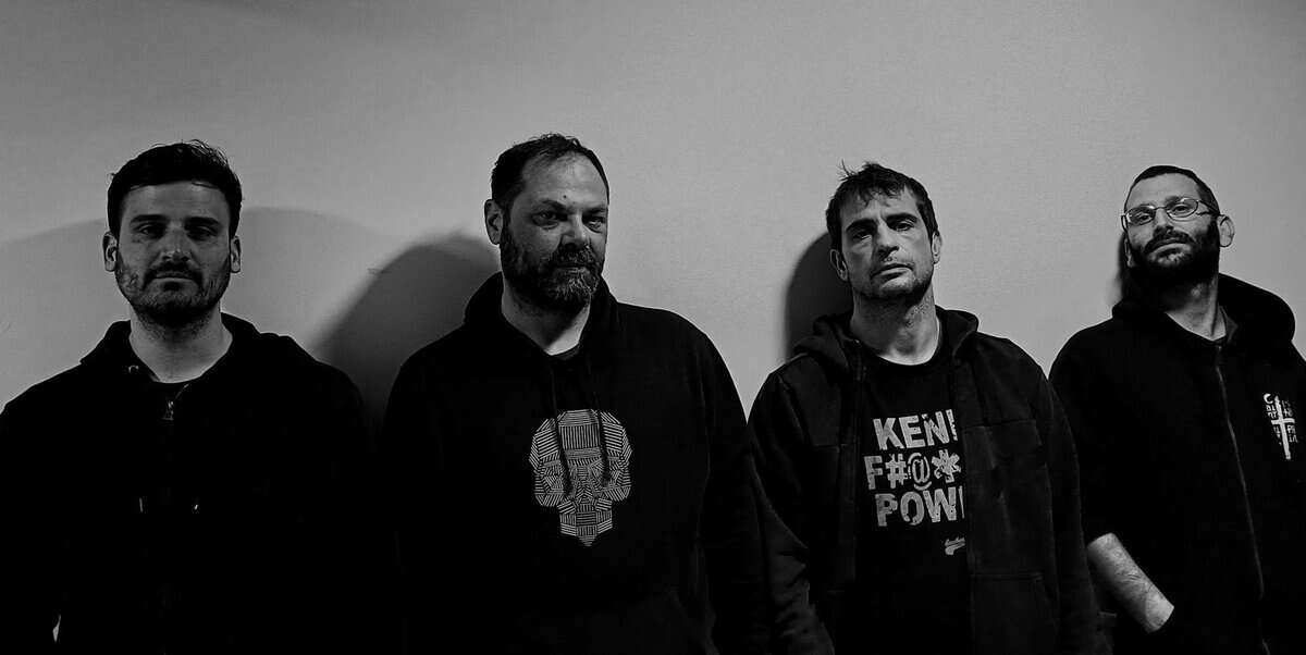 Four men in dark hoodies stand in a line against a plain wall, looking toward the camera in a black-and-white photo (band photo).