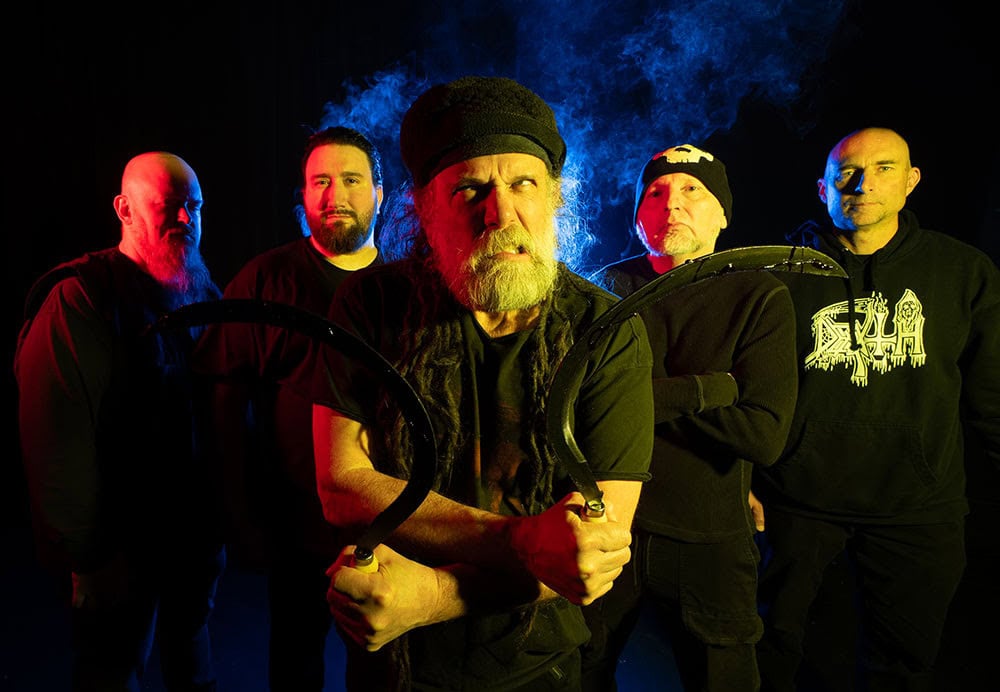 Five men in black clothing pose in a dark studio with dramatic blue and red lighting; the central bearded man grips two crescent sickles and stares forward.
