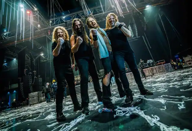 Four rock musicians pose together on a dimly lit stage after a concert, thumbs up and smiles, with streamers on the floor and equipment in the background.