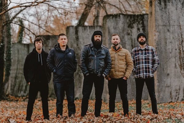 Five men standing in a line outdoors among autumn leaves, wearing jackets, posing for a group photo.