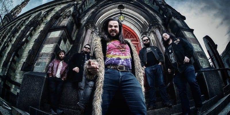 Five-man rock band posing on church steps, lead singer in a colorful tie-dye shirt and fur coat, shot from a low angle outside a Gothic building