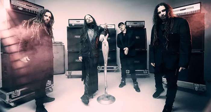 Four rock band members pose in a studio surrounded by large guitar amplifiers behind them.