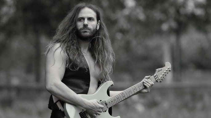 Guitarist with long hair and a sleeveless vest playing a white electric guitar outdoors.