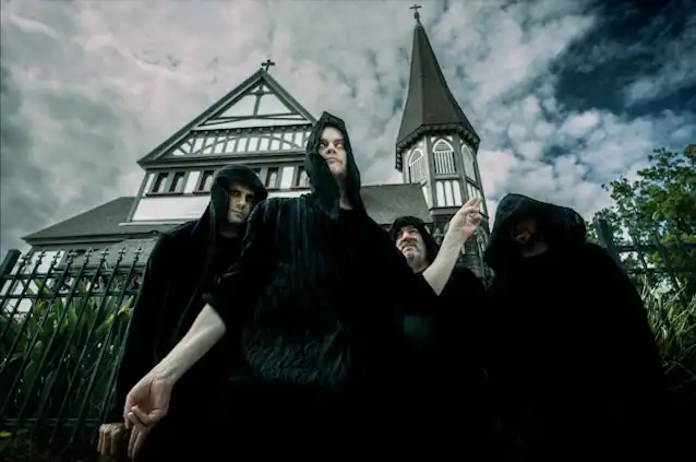 Four hooded figures in black cloaks stand outside a Gothic church with a tall steeple.