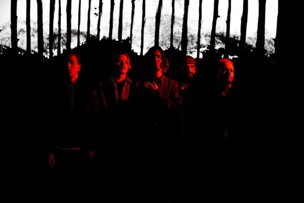 Five people standing in a dark venue with red lighting, silhouetted against a row of vertical bars.