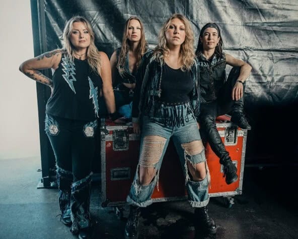 Four female rock musicians pose confidently in front of a red equipment case, wearing distressed jeans and black leather outfits.
