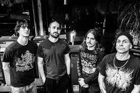 Four male rock musicians standing side by side backstage with amplifiers in the background.