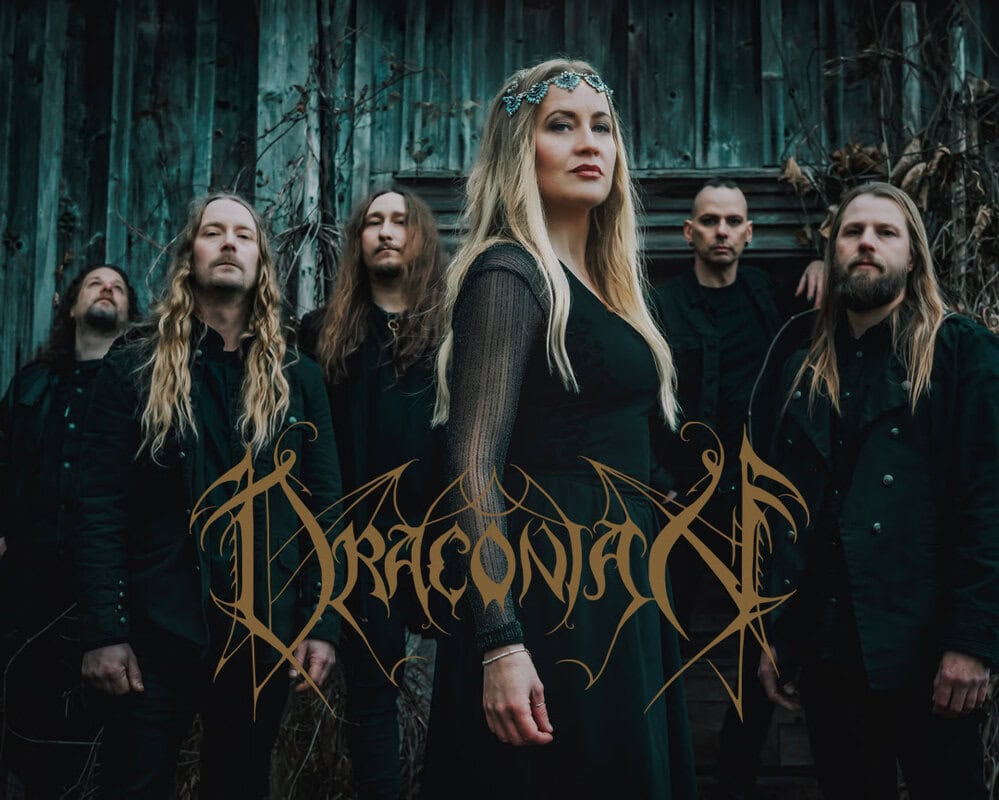 Female lead singer in a black dress with a jeweled headpiece stands front and center while five band members in dark clothing pose behind her in a moody, outdoor setting with a wooden wall behind.