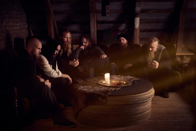 A group of men sits in a dim, rustic attic around a round table with a lit candle in the center.