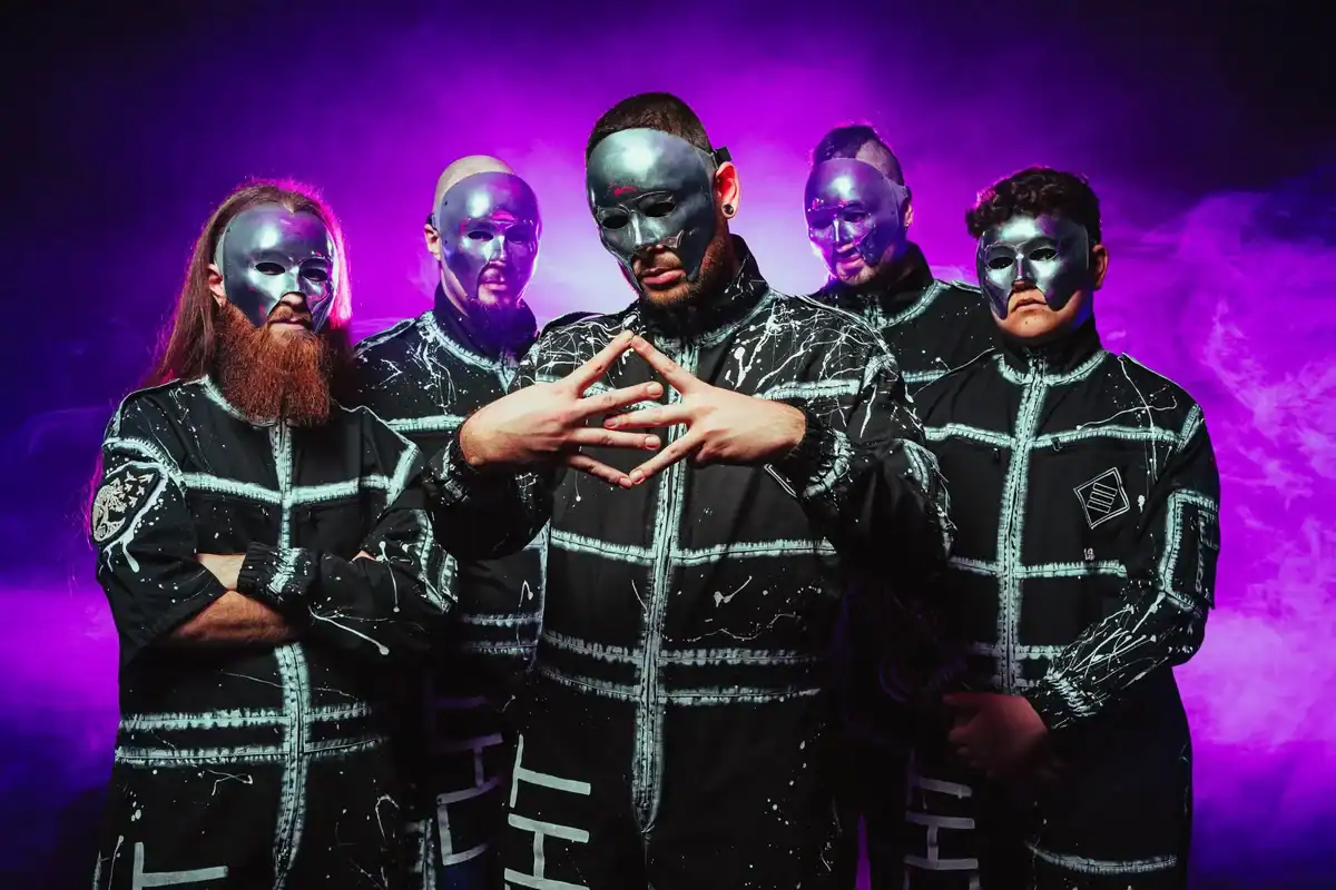 Five performers with silver masks and black splattered outfits pose against a purple smoky background.