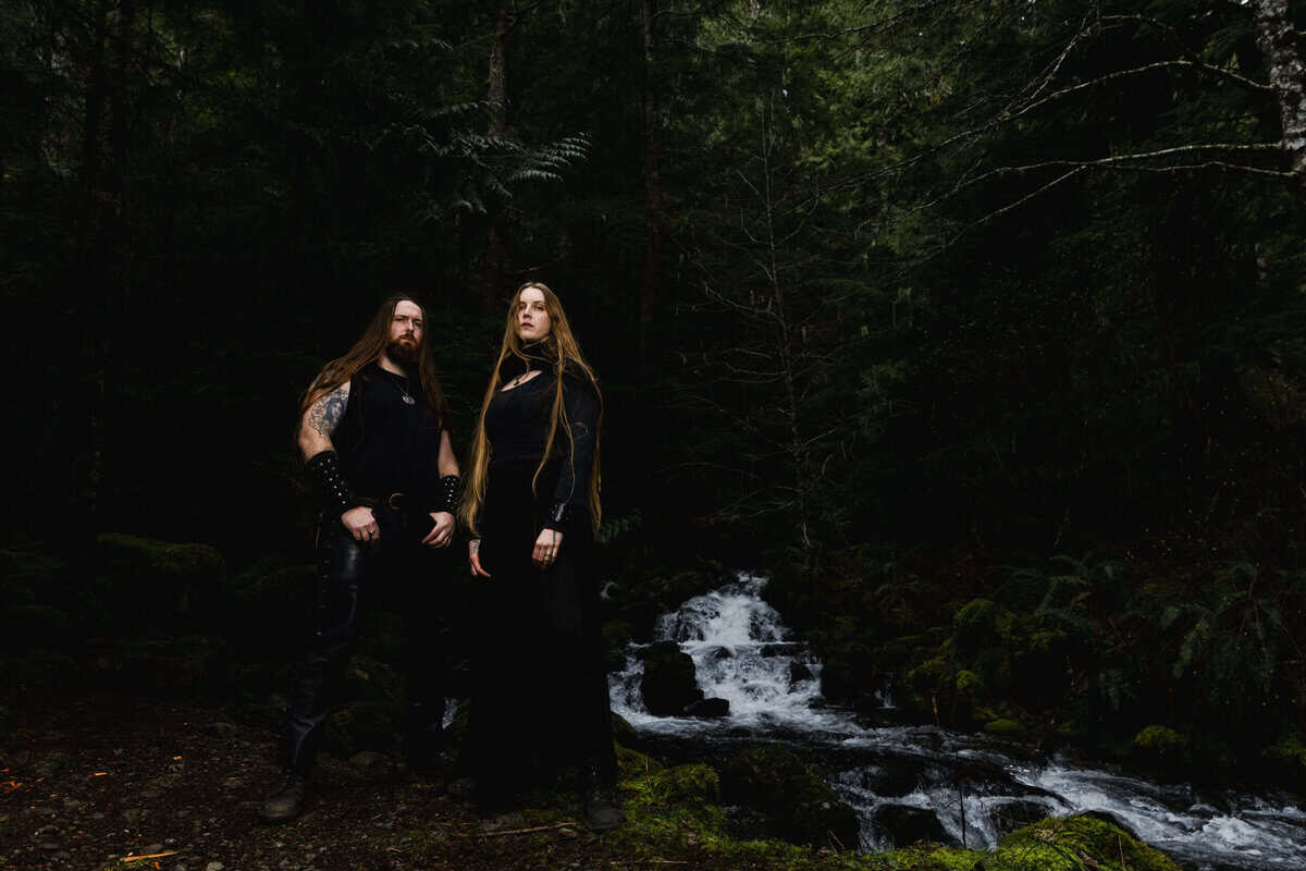 Two people with long hair in black clothing stand by a mossy forest stream.