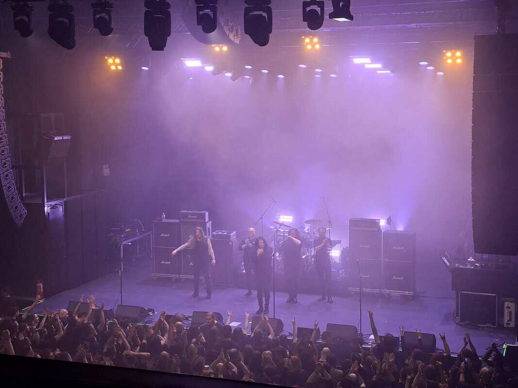 Live concert scene: performers on a purple-lit stage with fog and amplifiers, crowd raising hands in front of them