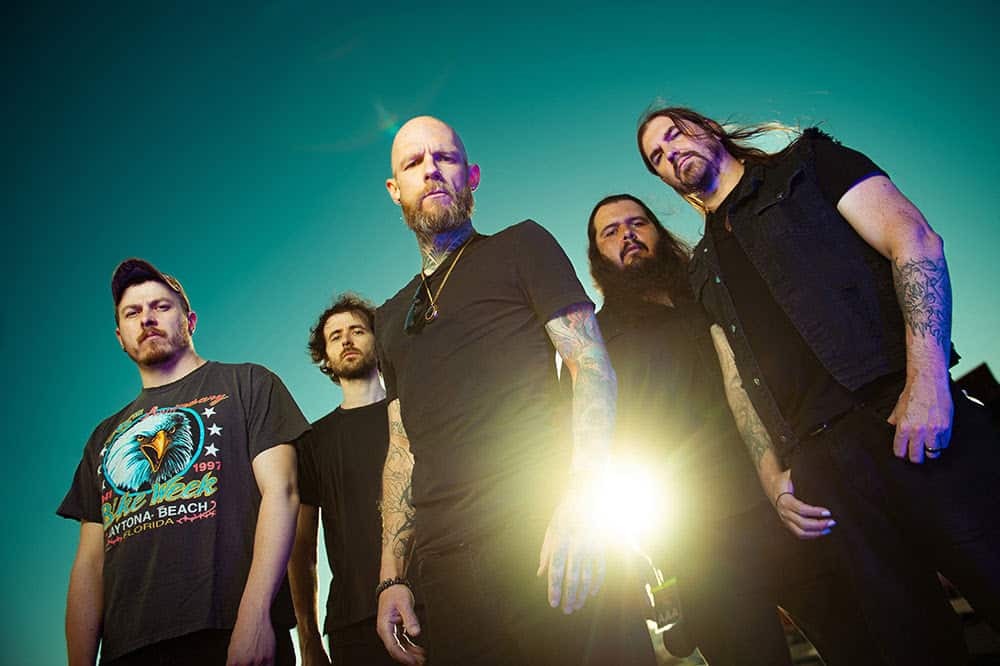 Five male rock band members pose together against a teal sky with a bright sun flare behind them, wearing dark clothing.