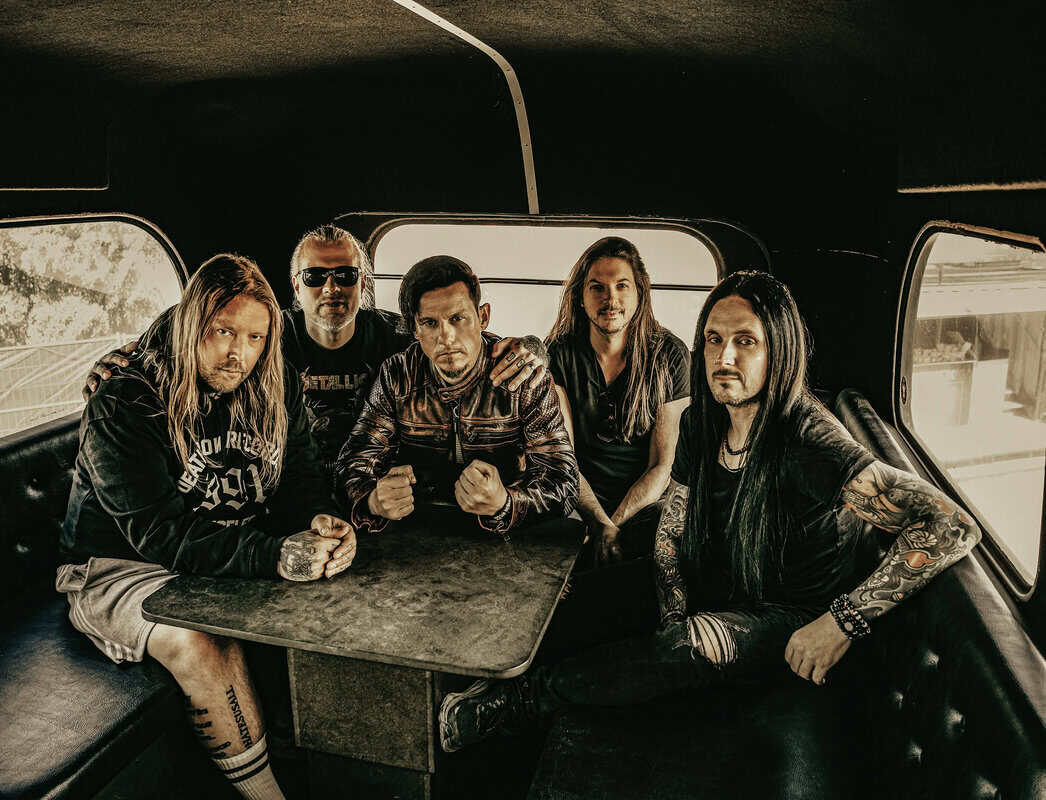 Five men with long hair and leather jackets sit close together inside a vehicle, posing for a band photo in a dim van interior.