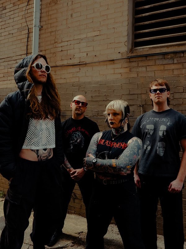 Four people in dark, edgy outfits pose against a brick wall; one with oversized hood and round sunglasses, others with tattoos and bold makeup, forming a punk-style group photo.