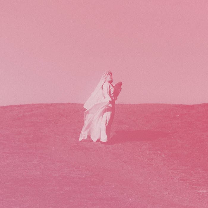 Bride in a long white wedding dress with veil walking on a grassy hill, carrying a child