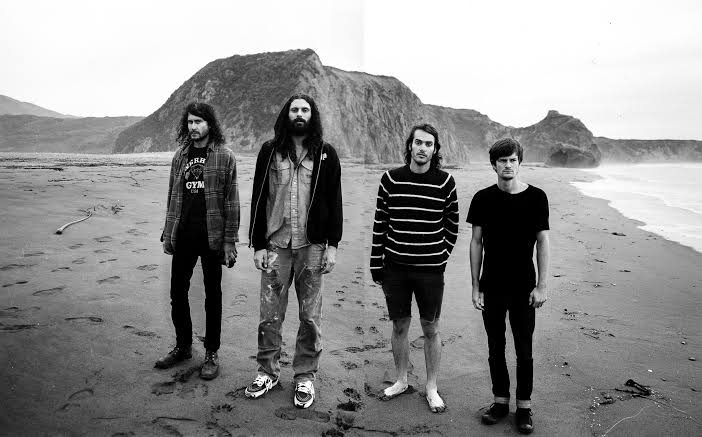 Four men stand on a foggy beach with large rock formations in the background.
