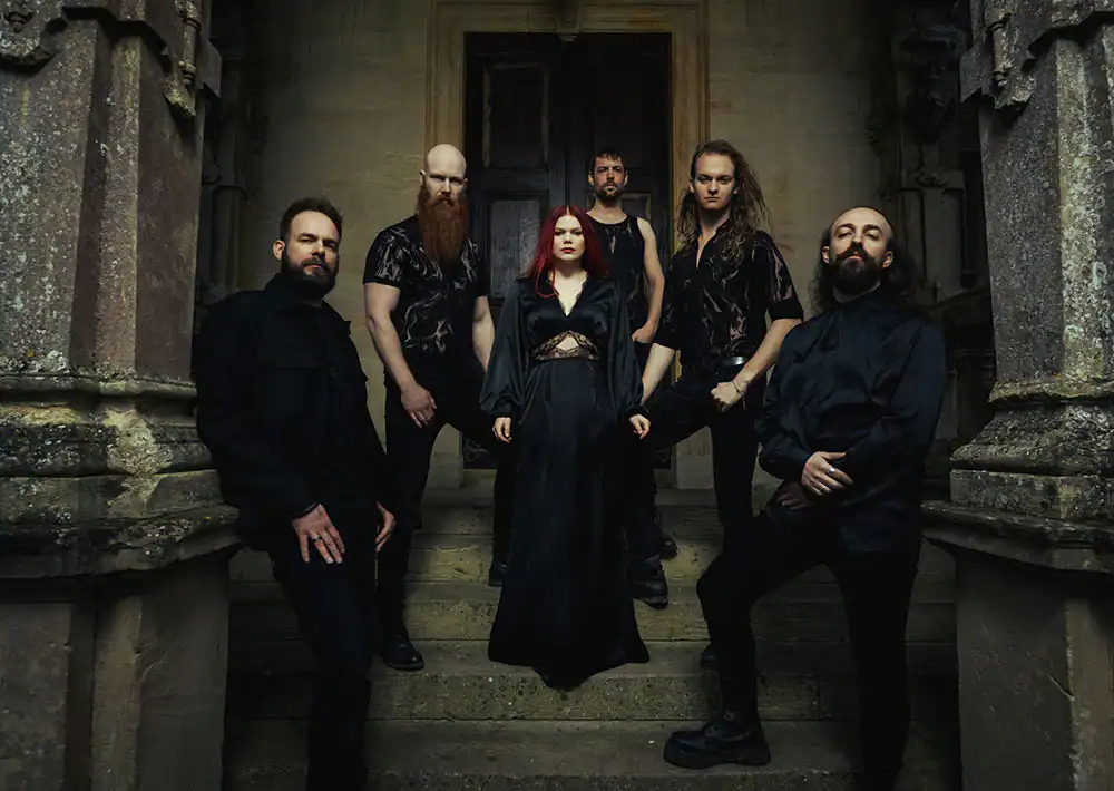 Six-member metal band posing on stone steps outside a weathered building in black attire, centered by a female vocalist with red hair in a cloak.