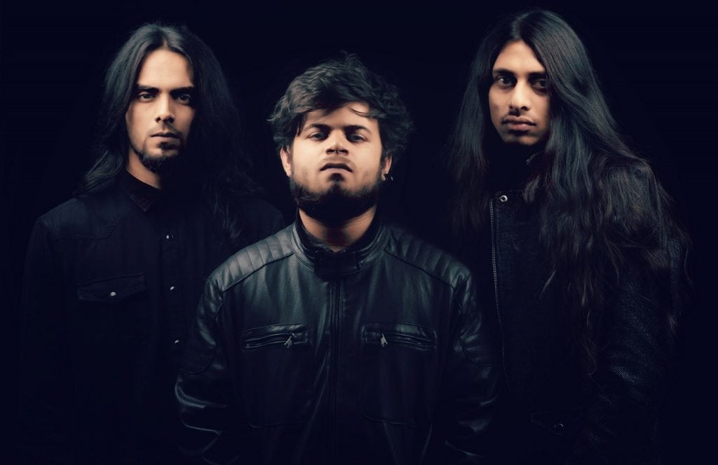 Three men with long dark hair posing in a studio against a black background; center man wears a black leather jacket and faces the camera.