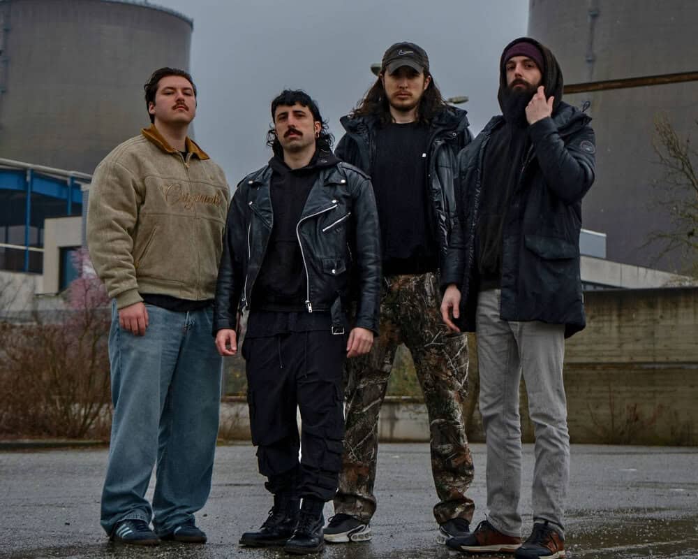 Four men pose together in an industrial outdoor setting, wearing jackets and casual clothing under a gray sky.