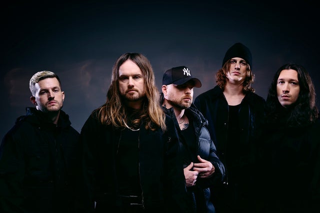 Five-member rock band posing in a dark studio, all wearing black jackets and moody lighting.
