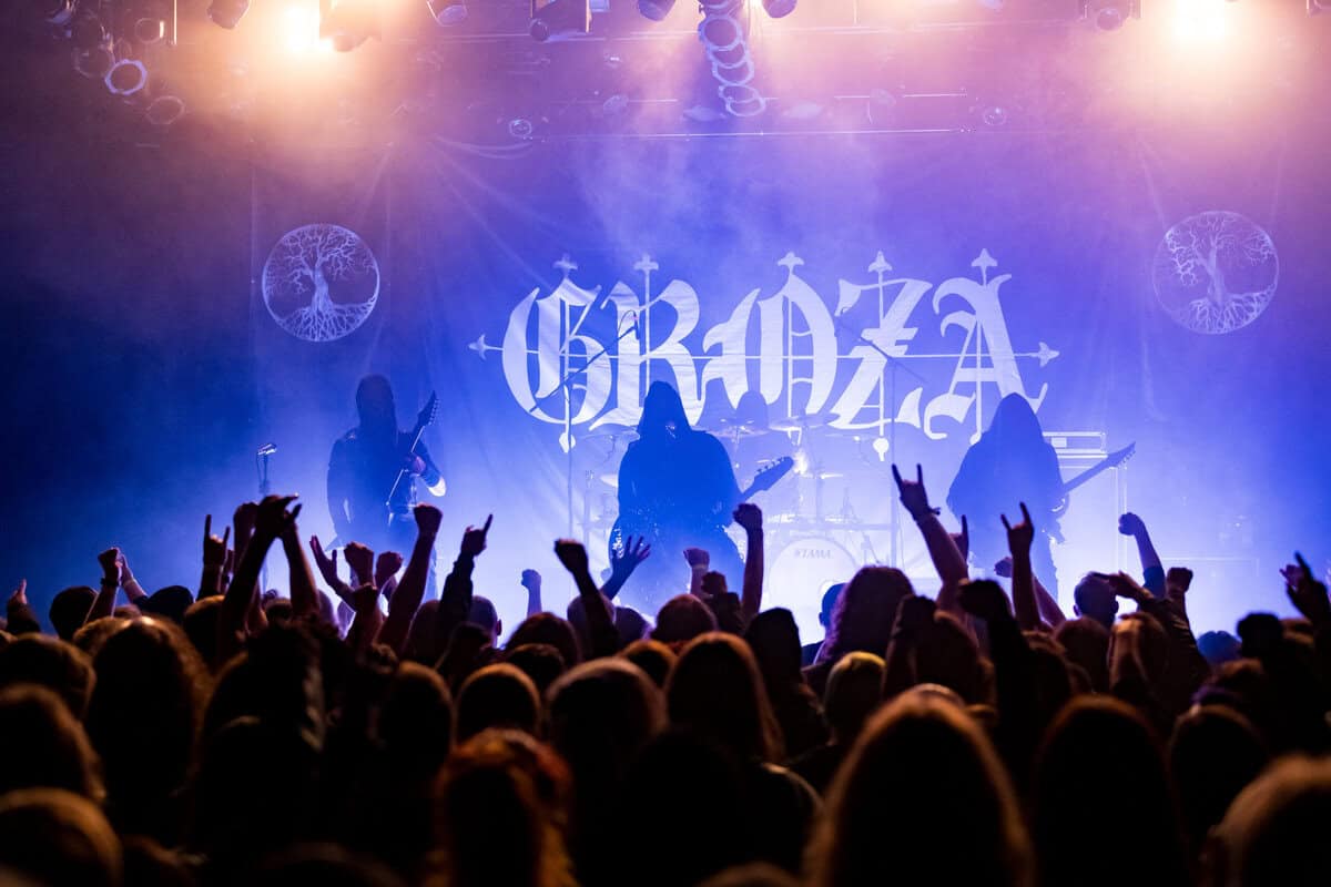 Band performing on stage with purple lighting, crowd raising fists, backdrop shows a large stylized logo with circular tree symbols nearby.