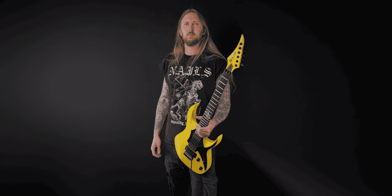 Man with long blond hair and tattoos holds a bright yellow electric guitar in front of a dark studio backdrop, wearing a black band t-shirt.