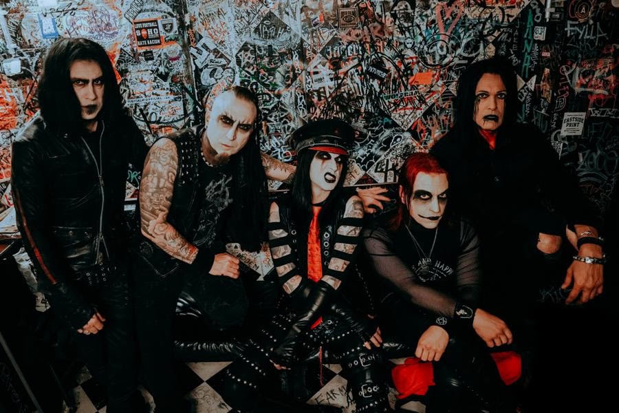 Five goth/metal band members pose together in a graffiti-covered backstage area, wearing black leather and heavy makeup for a dark, theatrical look.